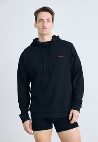 Mann iført en svart hettegenser med rødt "HUGO"-logo og svarte shorts, stående mot en lys ensfarget bakgrunn.