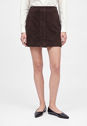 Femme portant un pull rayé blanc et noir, une jupe marron foncé, des ballerines noires pointues, avec de longs cheveux lisses et des boucles d'oreilles créoles, debout.