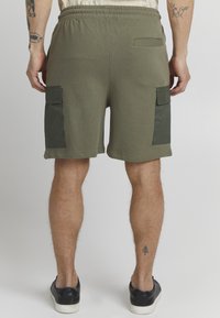 Shorts vert olive avec une taille élastique, deux poches cargo sur les côtés et une poche au dos, fabriqués en tissu doux, affichant un design décontracté.
