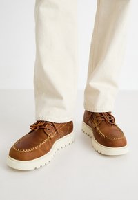 Hnědé kožené boty s bílou gumovou podrážkou, vybavené dekorativním prošíváním, kulatými tkaničkami a tradičním mokasínovým designem.