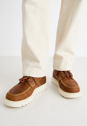 Chaussures en cuir marron avec une semelle en caoutchouc blanche, dotées de coutures décoratives, de lacets ronds et d'un design de mocassin traditionnel.