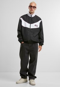 Veste coupe-vent zippée en noir et blanc avec des poignets élastiques, associée à un jean noir ample et des baskets crème. Accentuée par un logo brodé.