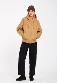 Tan capuchonjack met verstelbare trekkoord, ribgebreide manchetten en een voorzakken. Gecombineerd met zwarte wijde broek en zwarte sneakers.