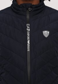 Mørkeblå polstret jakke med høy krage. Har diagonalt kviltet mønster, vertikal frontglidelås og EA7 Emporio Armani-logo på brystet.