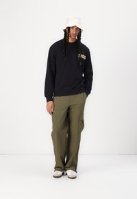 Zwarte sweatshirt met gouden tekst, olijfgroene cargobroek, witte schoenen met bruine accenten en een beige bucket hat. Casual, relaxte pasvorm.