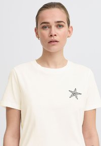 T-shirt en coton crème avec un graphique de étoile de mer noire sur la poitrine à gauche. Col rond classique avec des manches courtes. Texture lisse.