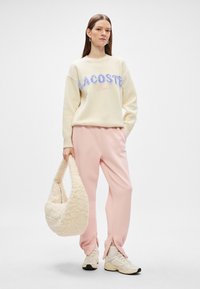 Κρέμα πλεχτό πουλόβερ με ανοιχτό μπλε και ροζ κεντητό λογότυπο "LACOSTE Paris", συνδυασμένο με μαλακά ροζ κολάν και μια αφράτη μπεζ τσάντα.
