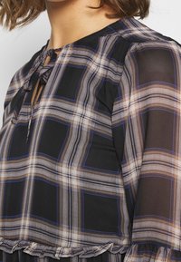 Musta sifonkiblouse, jossa on beige- ja sinivärinen ruutukuvio, avainreikäkaula-aukko ja frillahelma. Pehmeä rakenne ja laskeutuvat hihat.