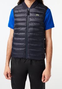 Námořnická péřová vesta s horizontálním prošíváním, předním zipem a kulatým límcem. Na levé straně hrudi je malá zelená značka.