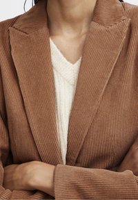 Brun cordflöjtsblazer med breda slag, vertikal ribbad textur och långa ärmar, lager-på-lager över en krämfärgad stickad tröja med V-hals.