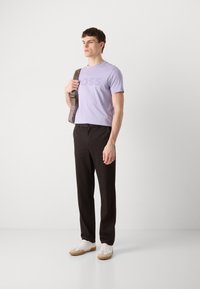 T-shirt violet clair avec le texte "BOSS", pantalon noir et baskets blanches avec des accents beige. Le jeune homme tient un sac à épaule.