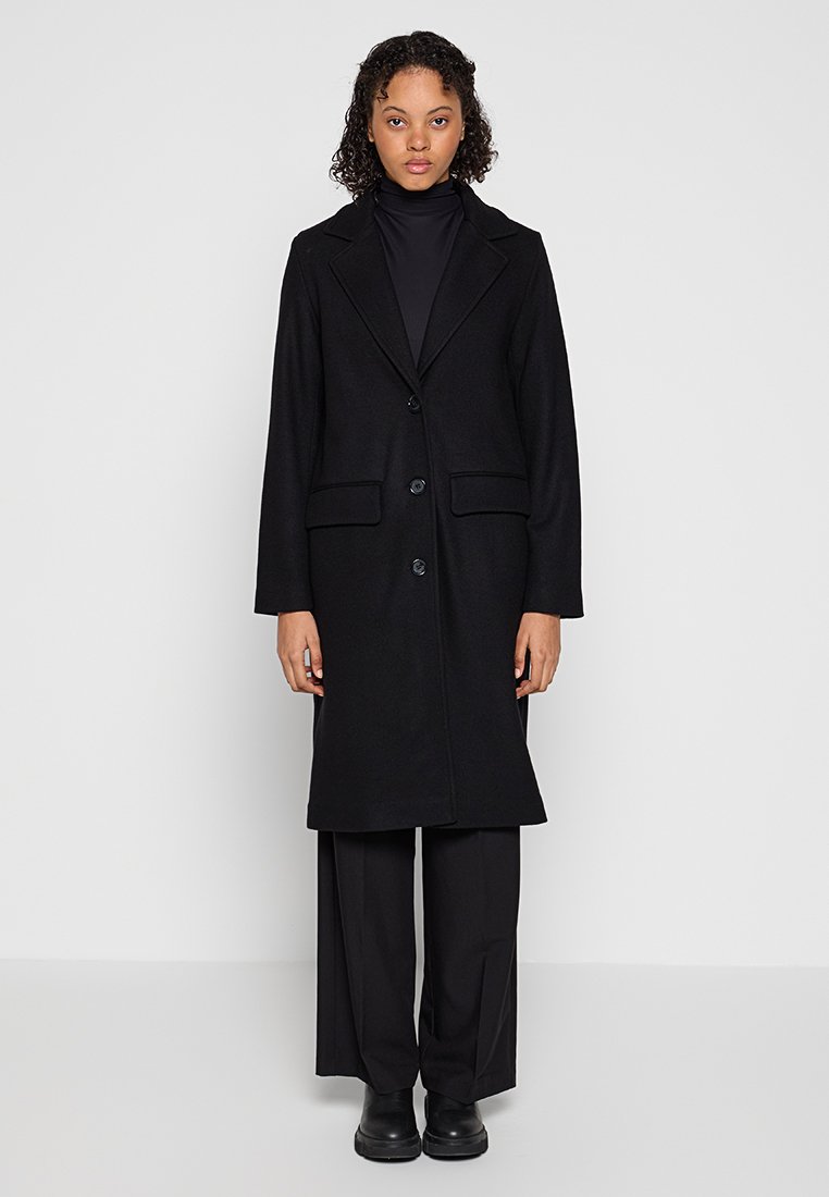Schwarz melierter Wollmantel mit maßgeschneidertem Design, langen Ärmeln, Reverskragen, vorderer Knopfverschluss und zwei seitlichen Taschen, getragen über schwarzen Hosen.