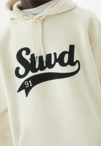 Haut à capuche couleur crème avec un logo "Stud" brodé en noir et le numéro "91" sur une bannière courbée. Comprend une capuche avec cordon de serrage et une poche avant.