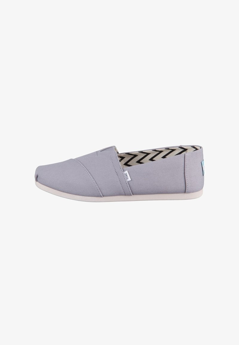 Zapato slip-on de lona gris con suela plana, que presenta costuras diagonales y un forro con patrón de chevron en blanco y negro.