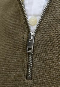Olīvzaļš zip-up džemperis ar teksturētu adījumu, ar sudraba metāla rāvējslēdzēju un baltu krekla apkakli redzamu zem tā.