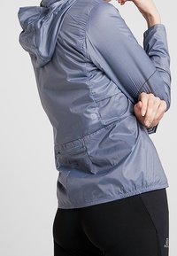 Veste légère et respirante de couleur bleue métallisée avec capuche, poignets élastiques, poches latérales et taille cintrée pour une silhouette ajustée.