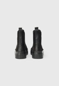 Černé semišové a kožené kotníkové boty s tlustými gumovými podrážkami a zadními poutky, zobrazené ze zadní strany na bílém pozadí.