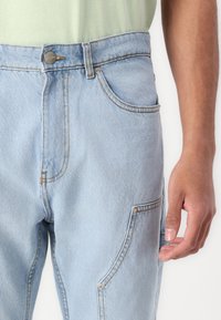 Light blue denim jeans featuring contrast stitching, a button closure, and multiple pockets, including a side pocket with snap closure.