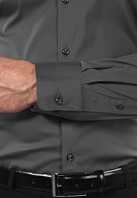 Camicia grigia con bottoni, caratterizzata da una texture liscia, bottoni neri e polsini ripiegati, abbinata a una cintura nera con finitura lucida.