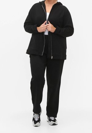 Person iført en sort lynlåshættetrøje, sorte bukser, hvid skjorte og sort-hvide sneakers, stående mod en ensfarvet hvid baggrund.