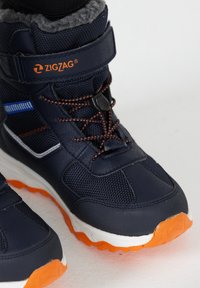 Dunkelblaue Winterstiefel mit orangener Sohle, strukturiertem Obermaterial, verstellbarem Riemen und Schnürsenkeln. Fleece-Futter am oberen Rand für Wärme. Zickzack-Logo sichtbar.