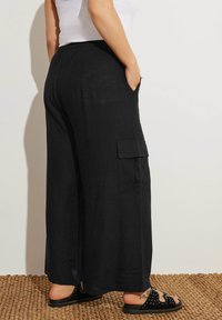Svarta linnebyxor med vida ben, sidofickor, elastisk midja och avslappnad passform. Bärs med platta svarta sandaler på en texturerad matta.