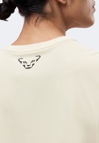 Κοντινό πλάνο από το άνω μέρος της πλάτης ενός ατόμου που φοράει ανοιχτό μπεζ πουκάμισο με μικρό μαύρο στυλιζαρισμένο λογότυπο ζώου κοντά στον λαιμό.