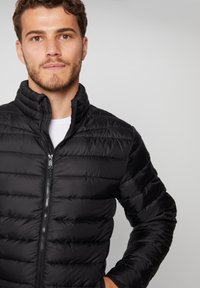 Chaqueta de plumas negra con un alto cuello, cierre de cremallera al frente y diseño acolchado. Material ligero con un acabado suave.