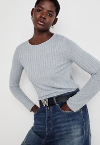 Lys grå ribbet langermet topp parret med blå denim jeans. Beltet har en sommerfuglformet spenne. Jeansen har en glatt tekstur og tettsittende passform.