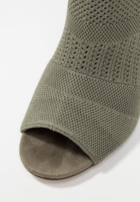 Talons ouverts de couleur vert olive avec un dessus texturé en tricot et une semelle intérieure douce, semblable à du daim. Présente un bord de pourpoint lisse et arrondi.