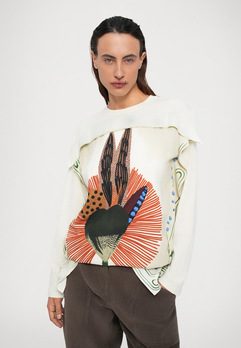 Una donna dai lunghi capelli scuri indossa una camicia a maniche lunghe color crema con una stampa astratta audace in arancione, blu e nero, e pantaloni di velluto a coste marrone scuro, con le mani in tasca.