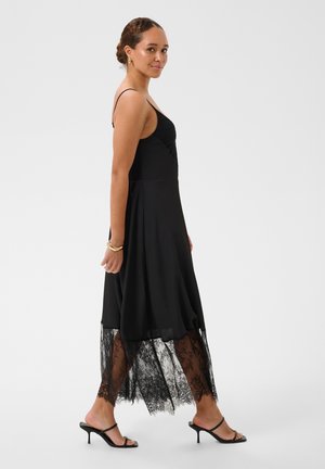 Femme en robe noire sans manches avec un ourlet en dentelle et des sandales noires à talons marchant devant un fond gris uni.