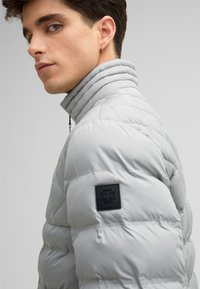 Giacca imbottita grigio chiaro con colletto alto, motivo trapuntato e una patch logo nera sul manica. Texture morbida e forma affusolata.