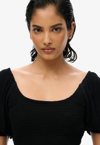 Sort struktureret top med firkantet halsudskæring og puffede ærmer. Stoffet er glat og har et ribbet design. Håret er slicket tilbage, neutral baggrund.