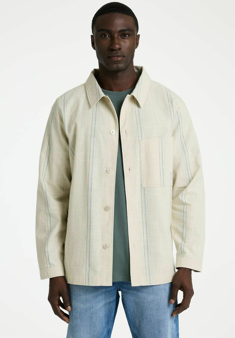 Hombre con chaqueta color crema abotonada con finas rayas verticales sobre camisa verde y vaqueros azul claro, posando frente a un fondo blanco.
