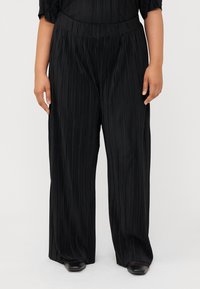 A person wearing black pleated wide-leg pants stands against a neutral background, showcasing the fabric's texture and style.