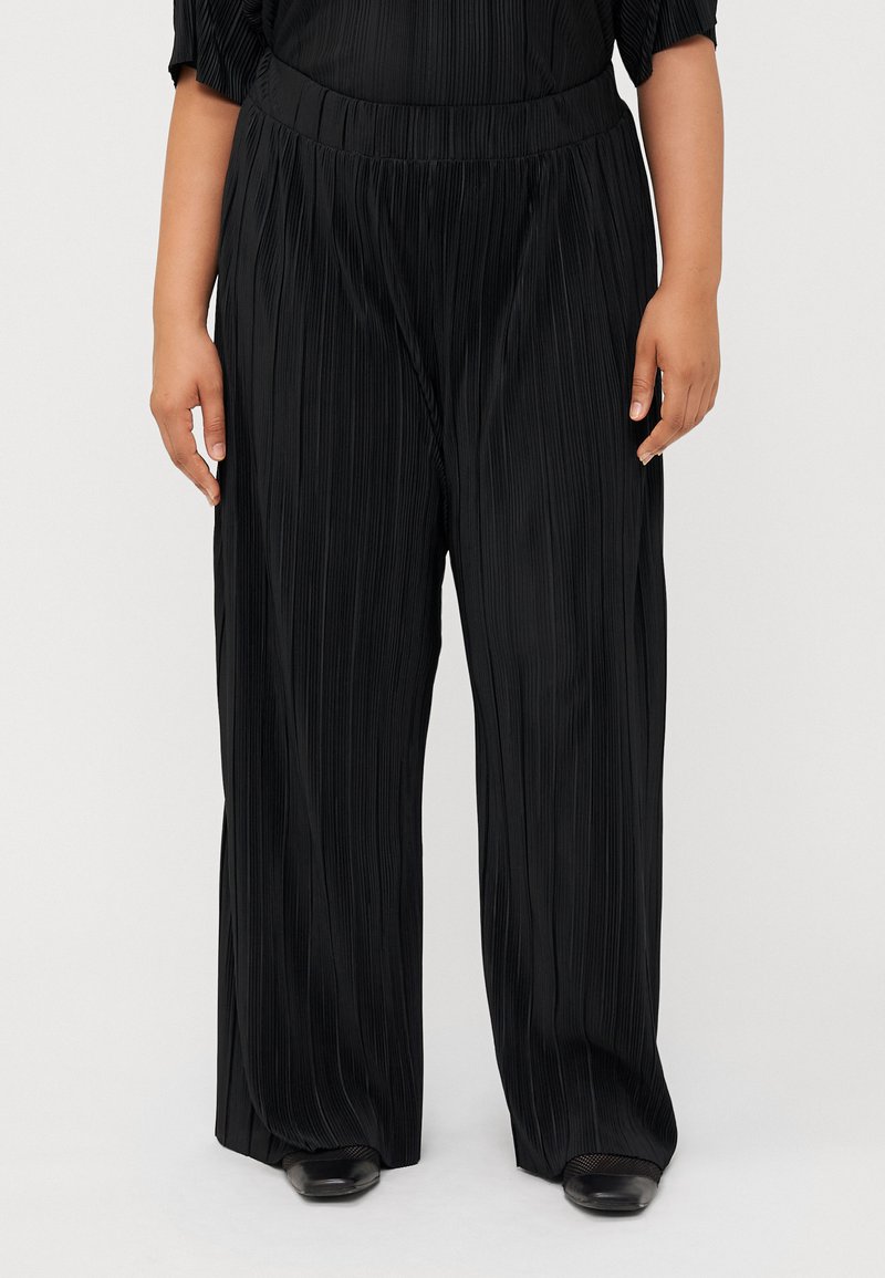 A person wearing black pleated wide-leg pants stands against a neutral background, showcasing the fabric's texture and style.