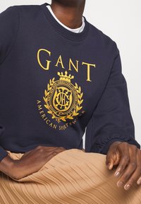 Felpa blu marina con stampa del logo dorato e design a corona. Presenta dettagli con polsini a coste. Indossata sopra una gonna beige a pieghe.