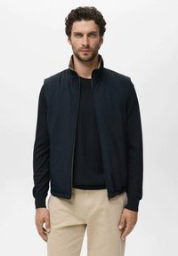 Marineblauwe mouwloze jas met een hoge kraag, ritssluiting en subtiele gouden accenten; gedragen over een zwart trui en beige broek. Gladde textuur.
