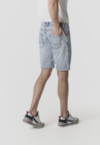 Homme portant un short en jean bleu clair et des baskets blanches, debout avec une main dans la poche arrière sur un fond uni.