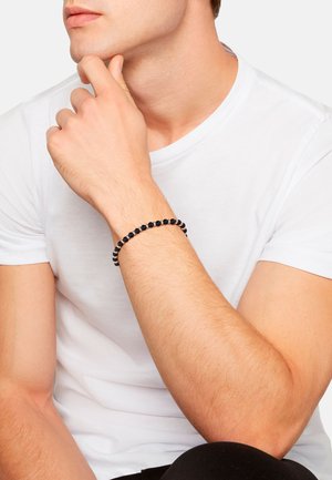 Man in white t-shirt resting chin on hand, wearing a black beaded bracelet on his wrist against a plain white background.