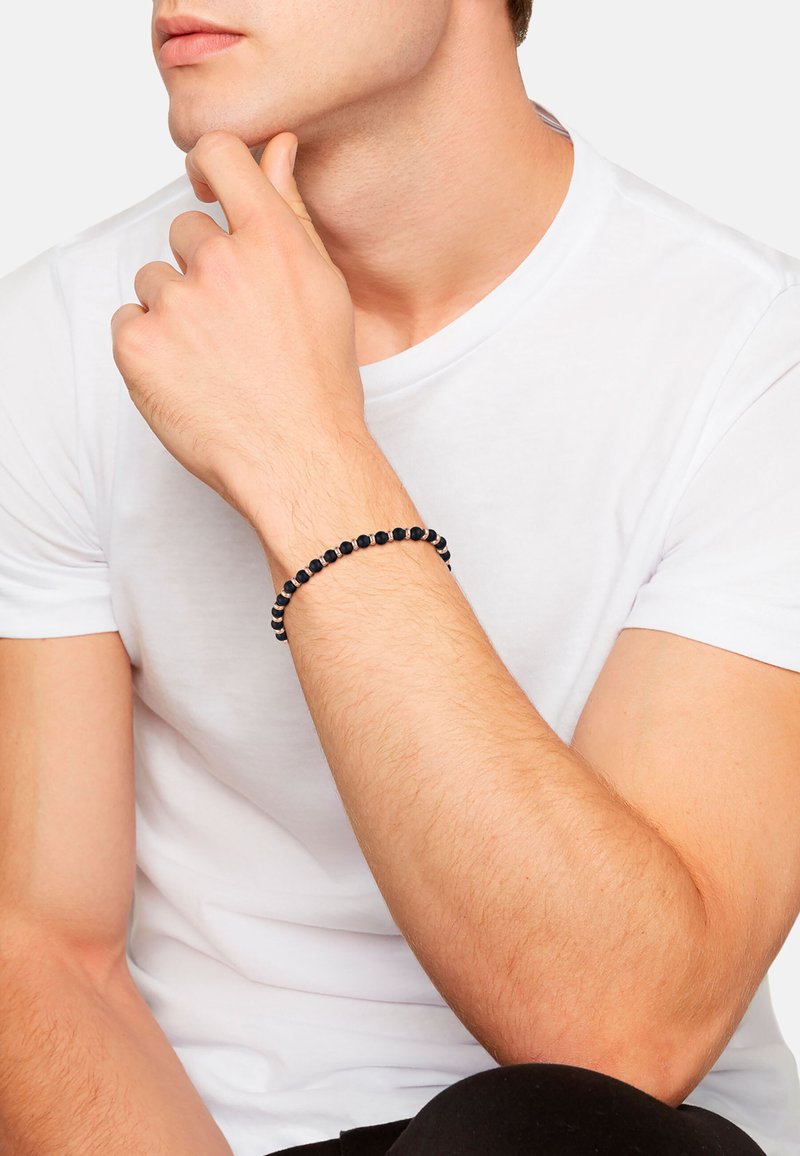 Muž v bílém tričku, který opírá bradu o ruku, na zápěstí má černý korálkový náramek, na jednoduchém bílém pozadí.