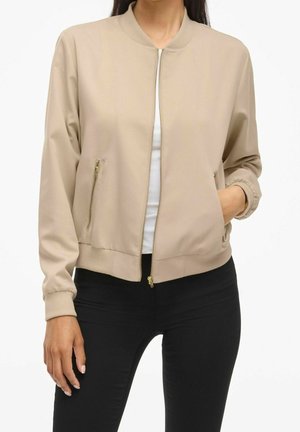 Vrouw draagt een beige bomberjack met rits aan de voorkant over een wit top en zwarte broek, met één hand in een jaszak.
