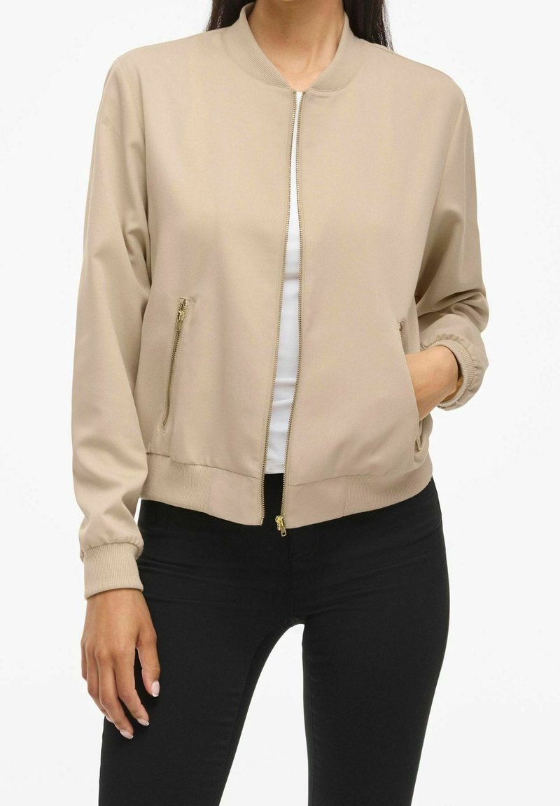 Femme portant une veste bomber beige zippée sur un haut blanc et un pantalon noir, avec une main dans la poche de la veste.