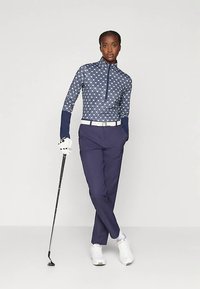 Nő áll keresztbe tett lábakkal, golfütőt tartva, mintás hosszú ujjú felsőt, sötétkék nadrágot, fehér övet, kesztyűt és sportcipőt viselve.