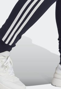 Marinblå mjukisbyxor med tre vita vertikala ränder; texturerat tyg vid fotmanschetterna; kombinerade med klumpiga vita träningsskor.