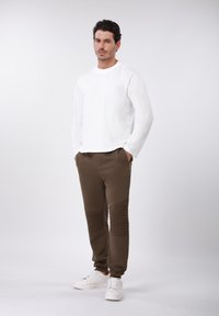 Λευκό πουκάμισο με μακριά μανίκια, καφέ παντελόνι με ριμπ υφή στα πόδια και λευκά αθλητικά παπούτσια, παρουσιασμένα μπροστά σε απλό φόντο.