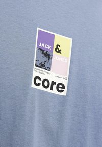 Ljusblå T-shirt med en rektangulär etikett som har "JACK & JONES" i svart och lila, och "core" i fet svart text. Mjuk textur.