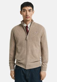Man in een beige zip-up sweater over een geruit shirt en een rode stropdas, gecombineerd met donkerblauwe jeans, tegen een eenvoudige witte achtergrond.