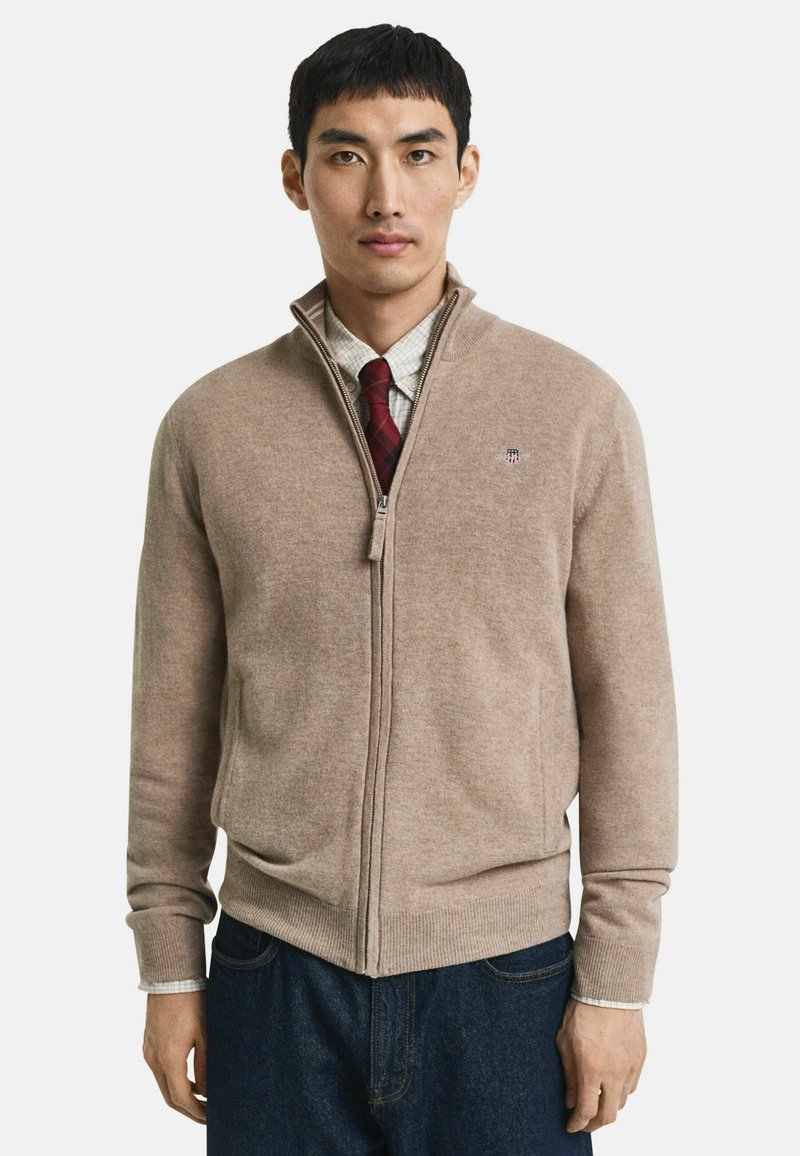 Man in een beige zip-up sweater over een geruit shirt en een rode stropdas, gecombineerd met donkerblauwe jeans, tegen een eenvoudige witte achtergrond.